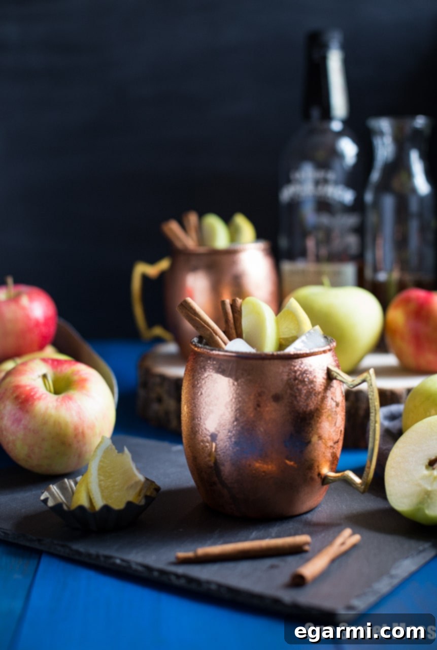 Apple Pie Mule in a copper mug, garnished with apple slices and cinnamon sticks, with autumn decor in the background.