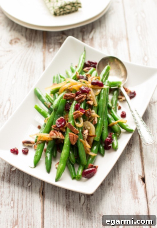 A festive Thanksgiving side dish: Crisp green beans with golden caramelized onions, toasted pecans, and ruby red dried cranberries in a white serving dish.